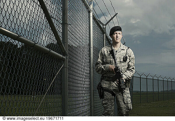 A Caucasian  male  Air Force Security Forces Airman in uniform poses with his M-4 rifle near the a secured fence line with barbed wire.