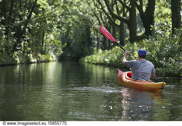 A caucasian man kayaking along a river shaded by a tree canopy