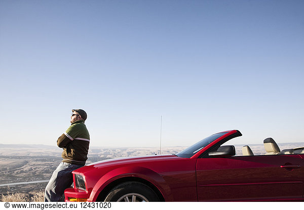A Caucasian man resting on the hood of his convertible sports car at a rest stop.