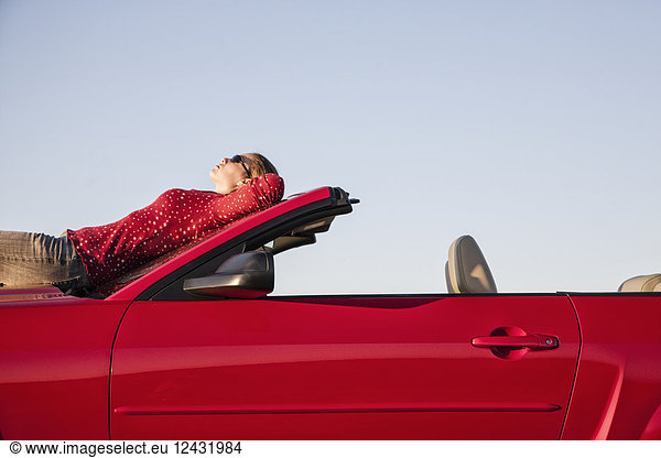A Caucasian woman resting on the hood of her convertible sports car at a rest stop.