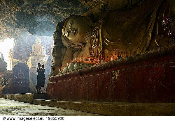 A cave temple  a large reclining Buddha statue  lit candles and a monk praying.