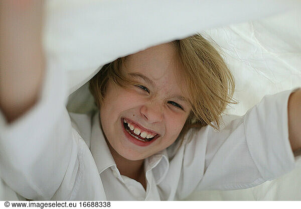 A child in a white shirt is fooling around under the covers.