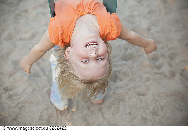 A Child Playing Outdoors. A Girl Hanging Upside Down On A Swing.