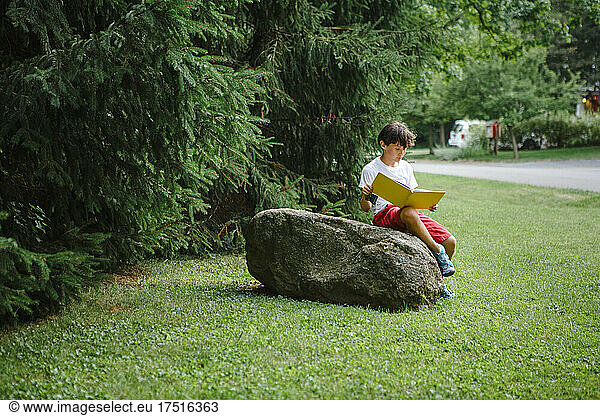 A child sits on boulder by side of road reading picture book