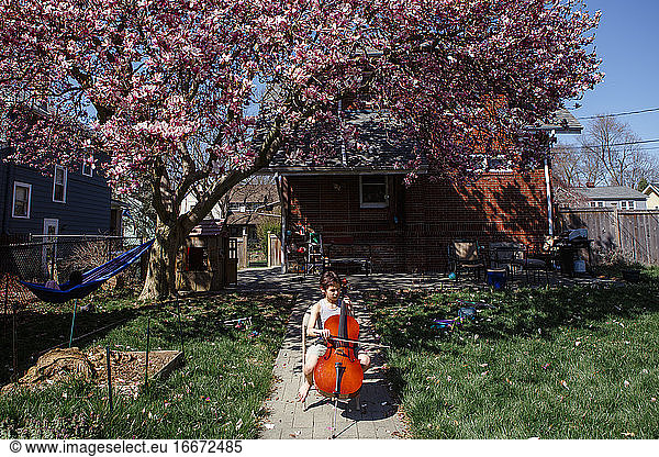 a child sits under flowering magnolia tree in backyard playing cello