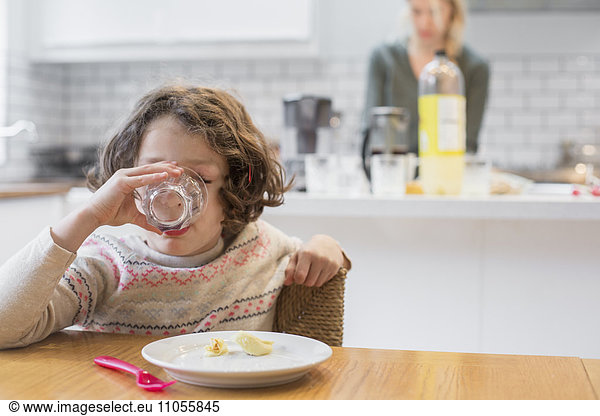 A child sitting at a table drinking from a glass  a woman standing behind her in a domestic kitchen.