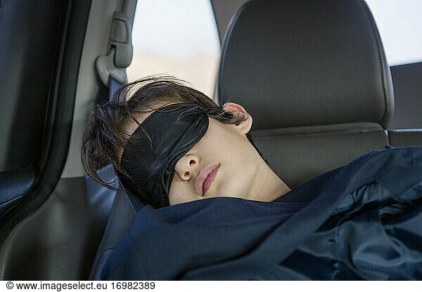 A child sleeps in a car seat wearing an eye mask covered in a blanket