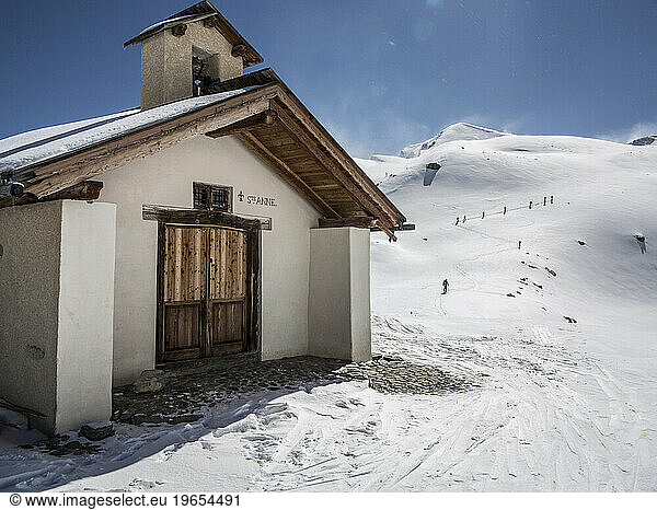 A church building in the mountains and skiers touring up a mountain in the background.