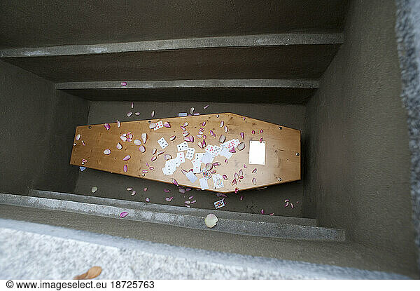 A coffin lays at the bottom of a grave with flower petals and paying cards