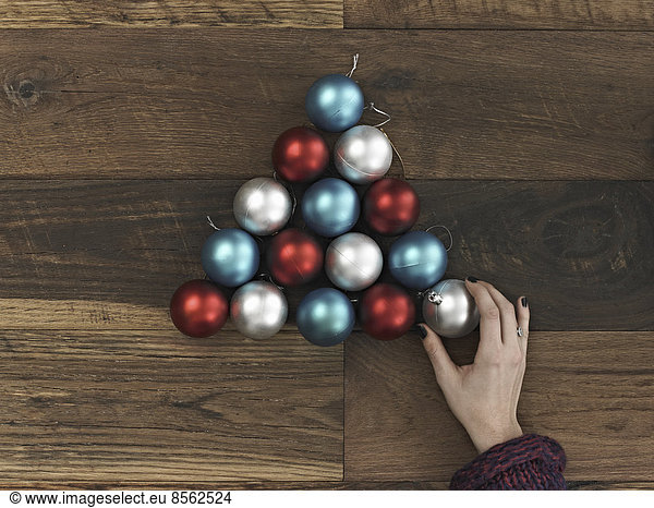 A collection of blue  red and silver ornaments arranged in a triangular shape on a wooden board. A Christmas tree shape. A person's hand placing the final ball.