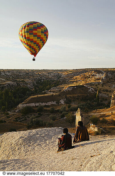 A Colourful Hot Air Balloon In Flight As Two People Sit On A Rock To Watch; Goreme  Cappadocia  Turkey