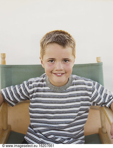 A confident boy sitting in a folding director's chair  smiling a toothy smile.