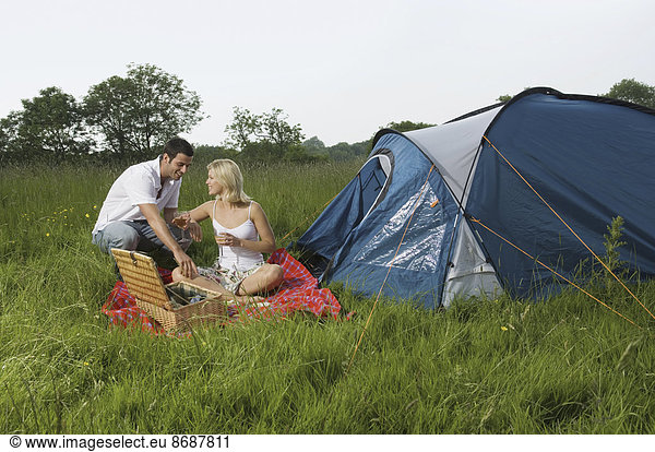 A couple  a man and woman seated having a picnic. A blue tent. Camping.