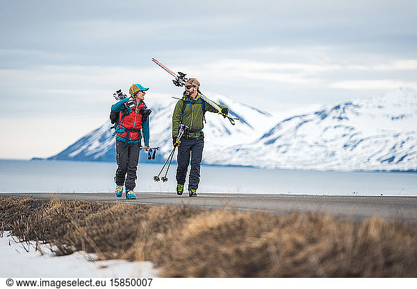 A couple walks down a road in Iceland holding ski gear