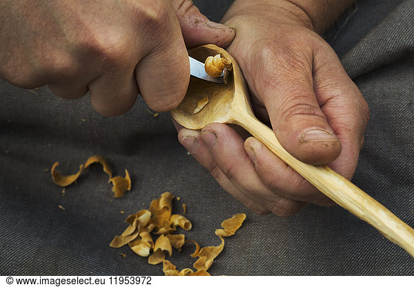 A craftsman carving wood  shaping the bowl of a handcarved wooden spoon with a sharp handheld tool.
