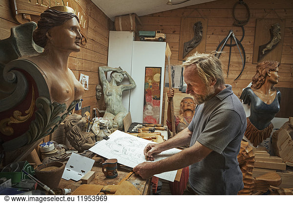 A craftsman  woodworker standing at a bench in a workshop working on a drawing  sketching using charcoal. Surrounded by carved wooden female ship's figureheads.