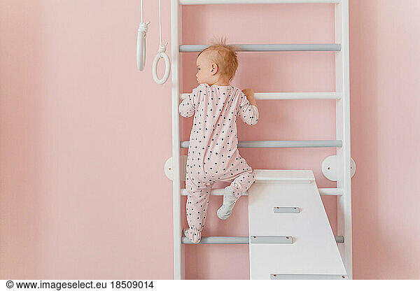 A cute toddler girl in soft pink pajamas does gymnastic on wall bars