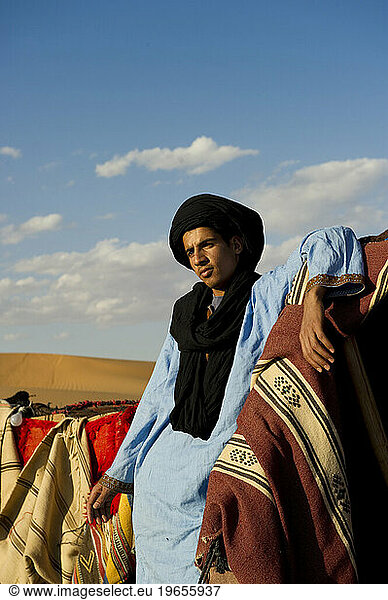 A desert guide in traditional dress stands in a tent camp with brightly colored blankets.