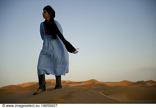 A desert guide in traditional dress stands on a dune at sunset.