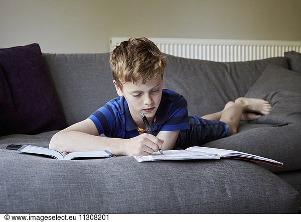 A family home. A boy lying on his front on a sofa doing his homework  writing in an exercise book.