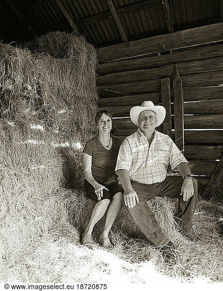 A farmer and his wife sit for a portrait on a bale of hay in a barn.