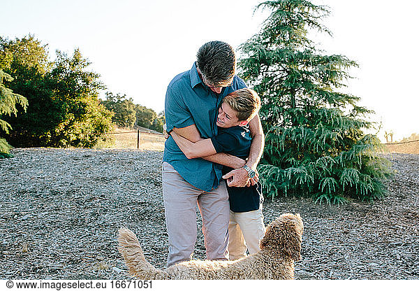 A Father And Teen Son Embrace While Outside With Their Labradoodle Dog