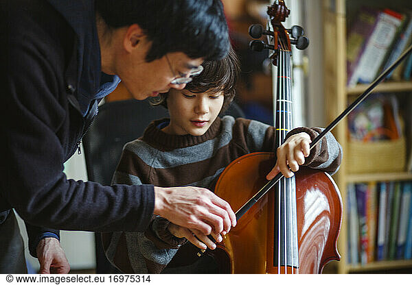 A father leans over child helping him learn to play a cello with a bow