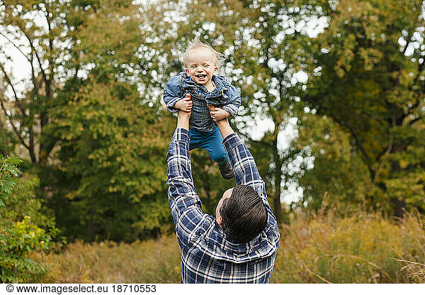 A father lifts his laughing baby boy high up into the air outside