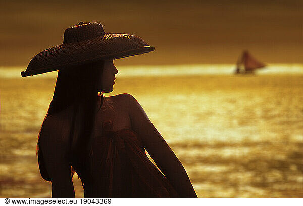 A Filipina wearing a traditional straw hat  in the distance  a sunset with an outrigger