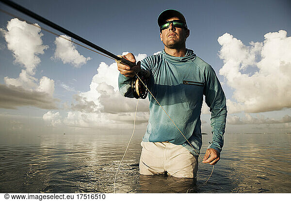 A fisherman in sunglasses pulls in fly line facing sun with clouds