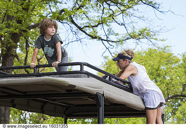 A five year old boy and a teenage girl on the observation platform of a safari jeep under trees.