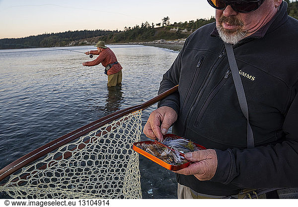 A fly-fishing guide checks his fly assortment for salmon and searun coastal cutthroat trout while his client fly fishes in the background.