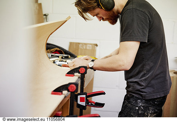 A furniture workshop making bespoke contemporary furniture pieces using traditional skills in modern design. A man at a workbench working on a piece of curved wood held with clamps.