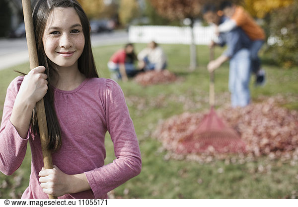 A girl holding a leaf rake and people raking fallen autumn leaves into piles.