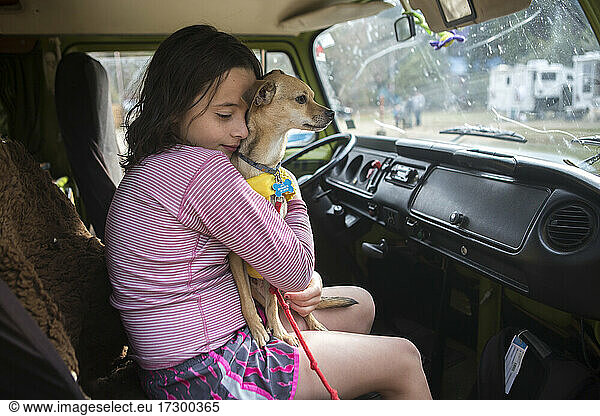 A girl hugs a chihuahua dog in VW camper van during roadtrip