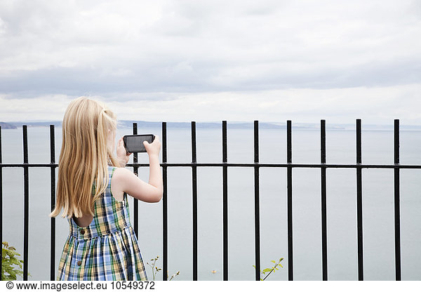 A girl in a checked sundress taking a photograph with a smart phone.