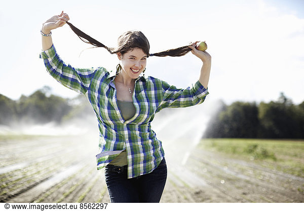 A girl in a green checked shirt with braids standing in a field with sprinklers working in the background.