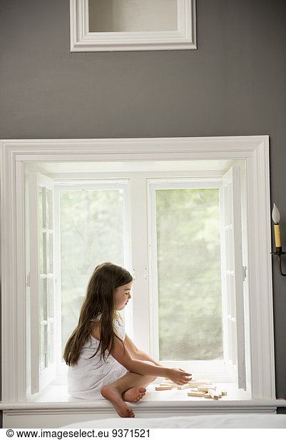 A girl in a white dress seated by a window playing.