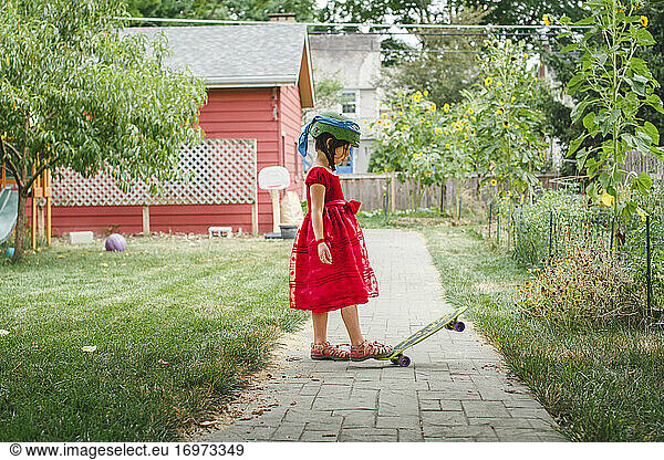 A girl in red dress and helmet steps on skateboard in garden in summer