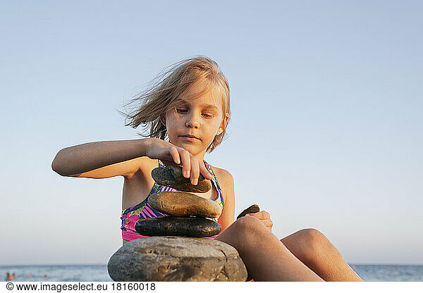 A girl sits on the seashore at sunset and plays with stones.