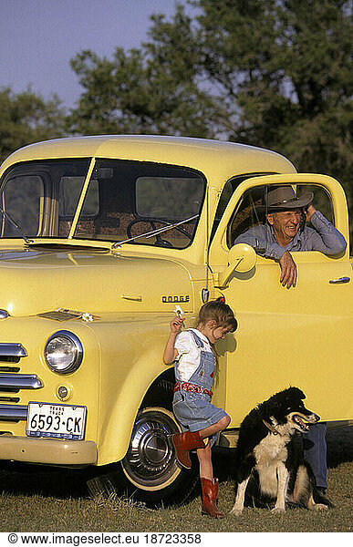 A grandfather takes his granddaughter and their dog out for a drive in an old yellow antique car.