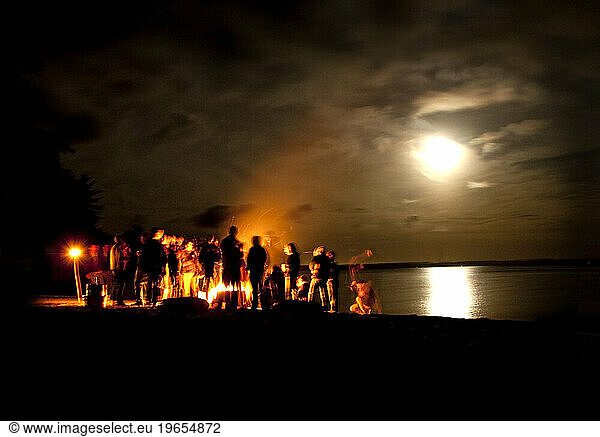 A group gathered around a campfire on the shore of Sebago lake.