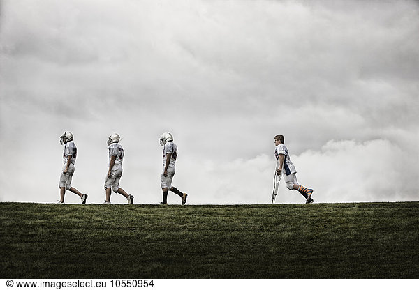 A group of football players  young people in sports uniform and protective helmets. Three fit players walking in line  one using crutches following.