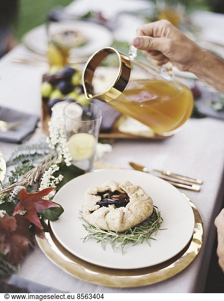 A group of people around a table in a garden. A celebration meal, with table settings and leafy decorations. A person pouring drinks into glasses.