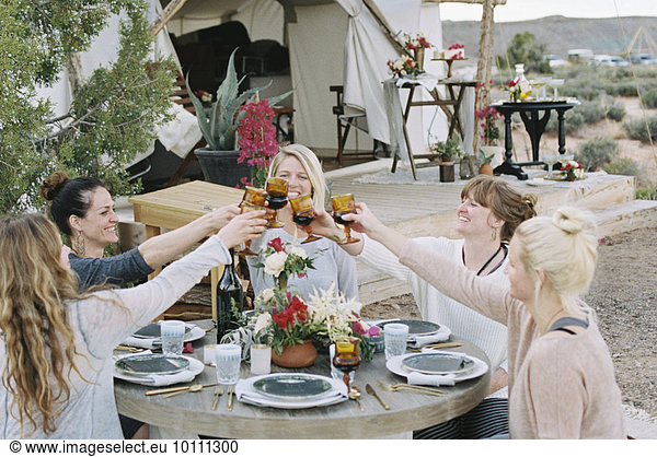 A group of women enjoying an outdoor meal by a large tent  in a desert landscape  raising a toast by clinking glasses.