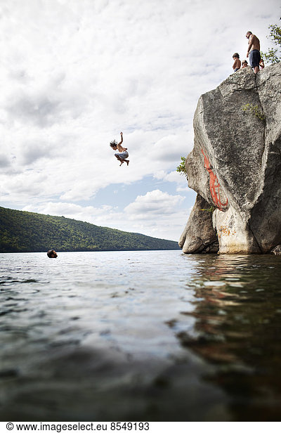 A group of young people jumping from a height from a cliff into the still waters of a lake.