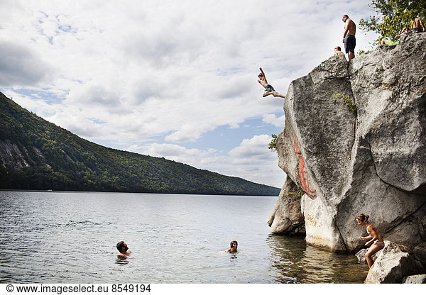 A group of young people jumping from a height from a cliff into the still waters of a lake.
