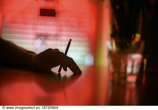 A hand holds a cigarette in a dimly lit bar.