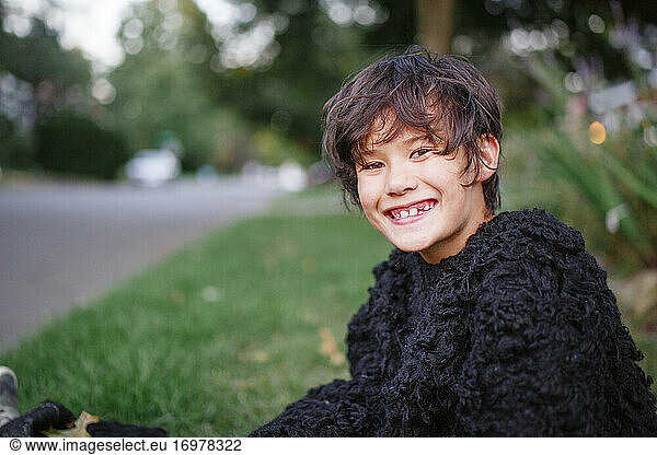 A happy boy in a gorilla suit smiles and sits in a grassy yard