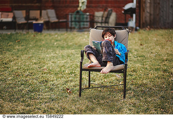 A happy boy reads a book barefoot in backyard in cool weather